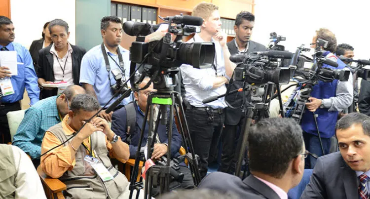 A group of reporters stand and sit around a row of large cameras on stands.  